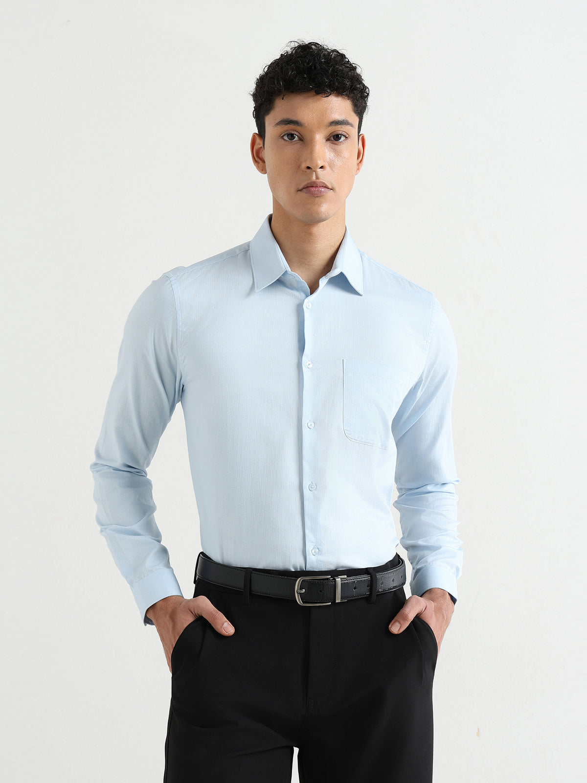 A man wearing a light blue long-sleeve dress shirt, black trousers, and a black belt, standing against a plain white studio background.