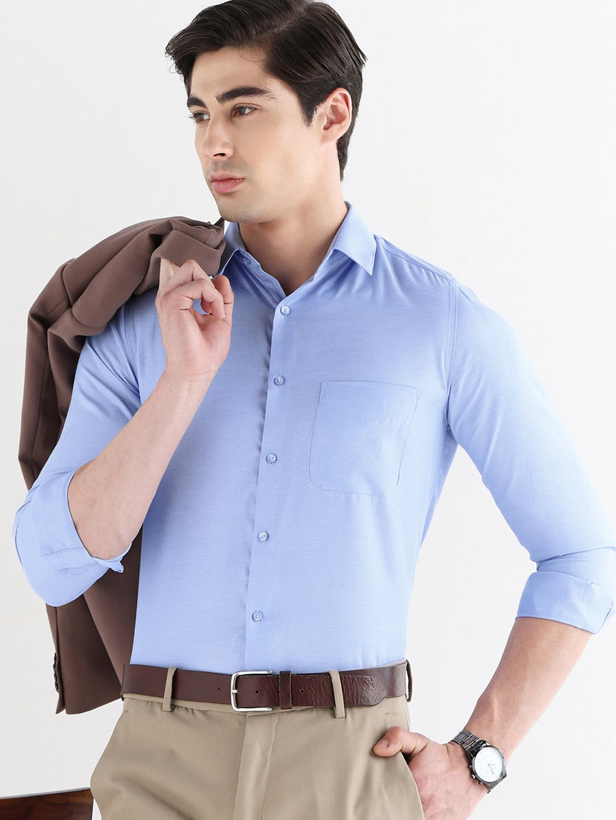 Man wearing a light blue dress shirt and beige pants, holding a brown jacket.