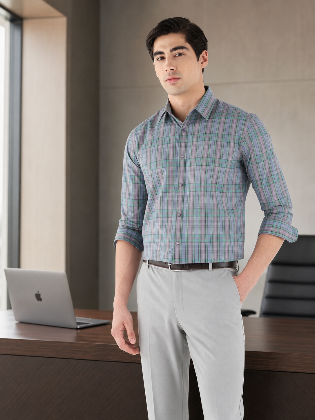 A man wearing a long-sleeve button-down plaid shirt in shades of blue, green, and gray, paired with light gray trousers, standing in a modern office environment next to a laptop on a desk.