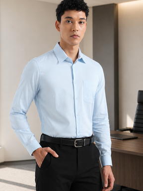 A man wearing a light blue long-sleeve button-down dress shirt, black trousers, and a black belt, standing in a modern office environment.