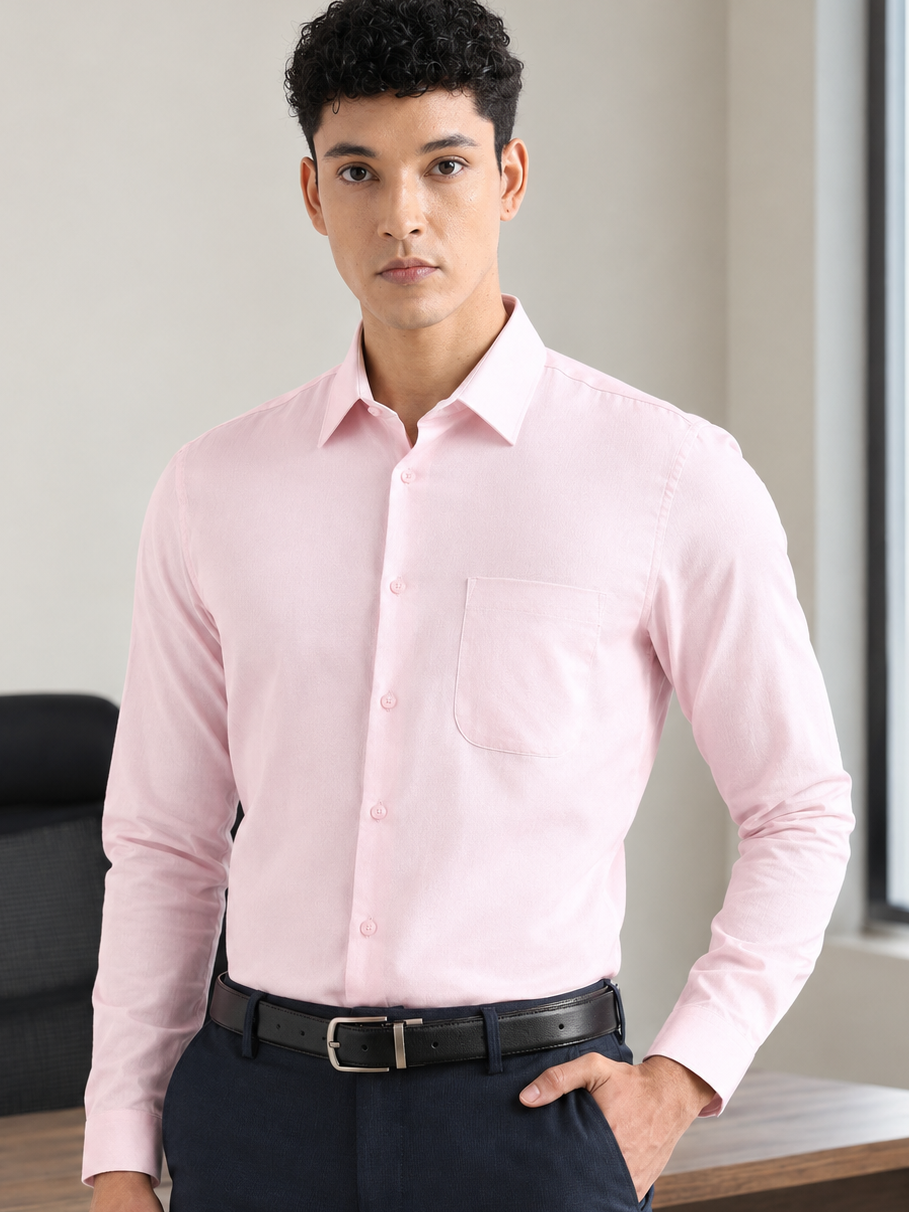 A man wearing a light pink long-sleeve button-down dress shirt, dark navy trousers, and a black belt, standing in an office setting.