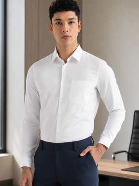 A man wearing a crisp white long-sleeve dress shirt and navy blue trousers, standing in a modern office setting with a window, desk, and chair visible.