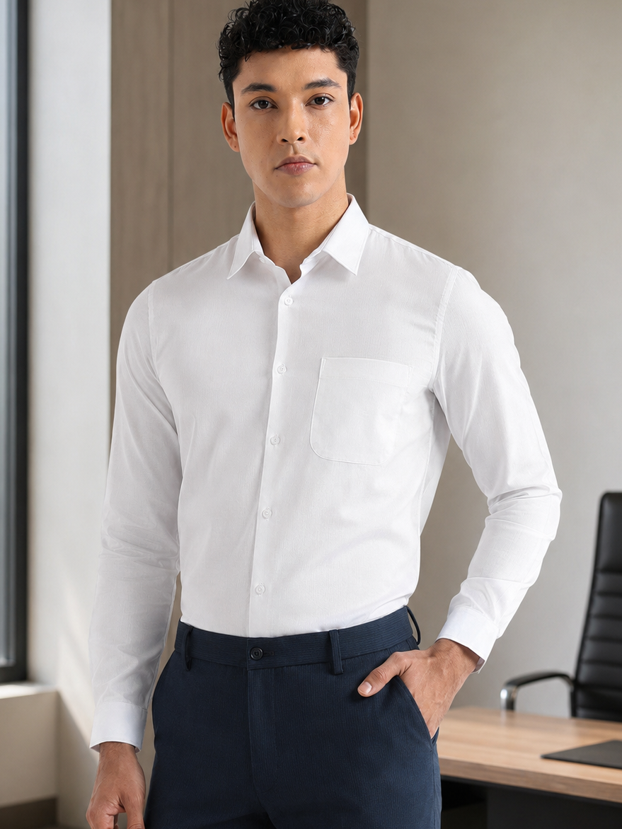 A man wearing a crisp white long-sleeve dress shirt and navy blue trousers, standing in a modern office setting with a window, desk, and chair visible.